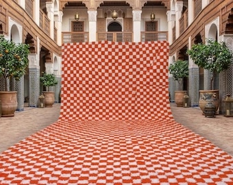 May include: A large, rectangular rug with a red and white checkered pattern. The rug is displayed in an outdoor courtyard setting with trees in large pots and decorative lanterns. The rug has a fringed edge.