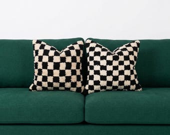 May include: Two square throw pillows featuring a black and white checkered pattern with a textured, tufted surface. The pillows are positioned side by side on a solid dark green sofa, showcasing a high-contrast geometric design.