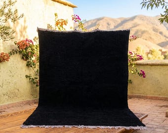 May include: A large, black rug with a textured surface and a white fringe. The rug is displayed outdoors against a backdrop of a mountain range and a building with flowering plants.
