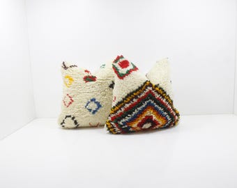 May include: Two cream-colored, textured throw pillows. One pillow features colorful geometric shapes, while the other has a vibrant, chevron-like pattern in red, yellow, blue, and black. These decorative pillows add a touch of bohemian flair.