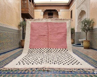 May include: A large, rectangular rug with a pink center, bordered by off-white and a lower section with black diamond patterns. The rug has a fringed edge and is displayed in a courtyard setting.