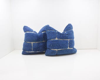 May include: Two blue decorative pillows with a textured, brick-like design. The pillows have a soft, plush appearance and are arranged side-by-side against a white background. The pillows are a vibrant blue color with white accents.