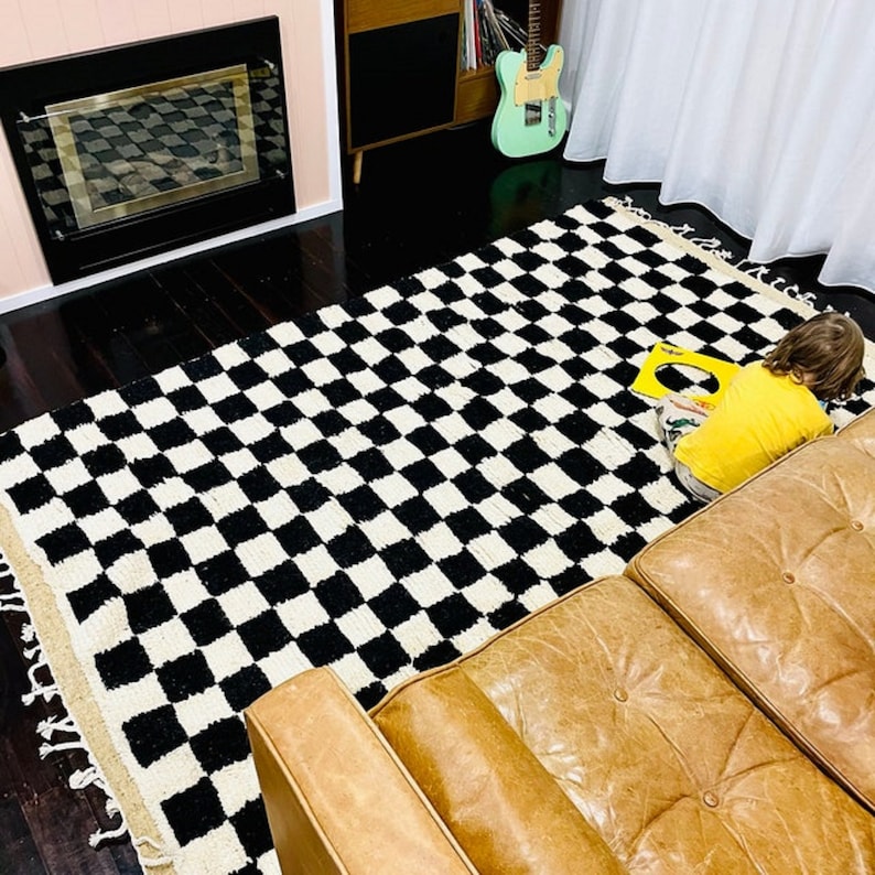 May include: A black and white checkered rug with fringe edges. A child is sitting on the rug playing with a toy. A brown leather couch is in the background.