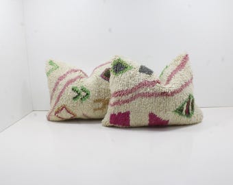 May include: Two textured throw pillows. The pillows are off-white with pink, green, and gray geometric patterns. The pillows are placed on a white surface, and the background is also white.