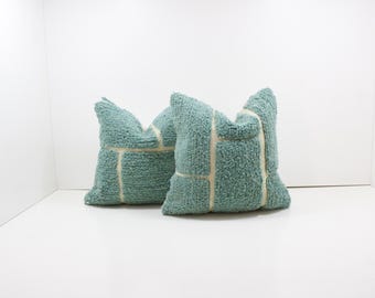 May include: Two square teal throw pillows with a textured, looped fabric. Each pillow features a cream-colored geometric design. The pillows are placed on a white surface against a white background.