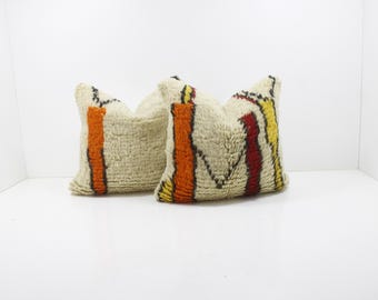 May include: Two beige throw pillows with vertical orange, red, and yellow stripes. The pillows have a textured, woven appearance and are placed against a white background. The pillows are decorative and suitable for a living room or bedroom.