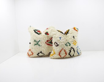 May include: Two cream-colored, textured throw pillows with colorful geometric patterns. The pillows have a shaggy, fluffy texture and are decorated with red, green, yellow, blue, and black shapes. They are placed against a white background.