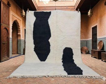 May include: A large, rectangular rug with a white background and two large, irregular black shapes. The rug has a thick, textured pile and a fringed edge. The rug is displayed in a room with arched doorways and ornate wooden doors.