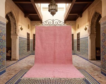 May include: A large, dusty rose-colored rug with a fringed edge is displayed in a hallway. The rug is the focal point, set against a backdrop of ornate architectural details, including tiled walls and arched doorways. The rug's texture is visible.