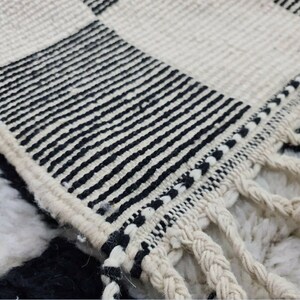 May include: Close-up of a black and white woven rug with fringe. The rug has a checkered pattern with black stripes.