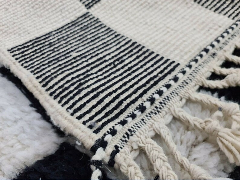 May include: Close-up of a black and white woven rug with fringe. The rug has a checkered pattern with black stripes.