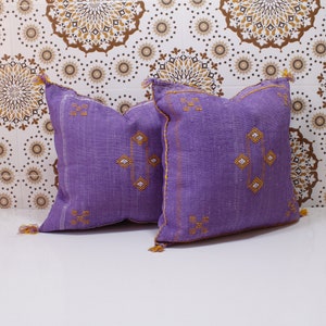 May include: Two purple throw cushions with embroidered geometric patterns in gold thread. The cushions are on a white surface with a brown and white patterned background.