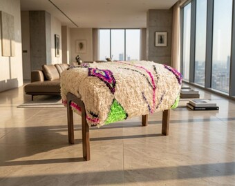 May include: A rectangular ottoman featuring a thick, cream-colored shaggy wool top with vibrant geometric patterns in neon pink, bright green, and black. The seat is supported by four dark brown wooden legs with a curved, minimalist frame design.