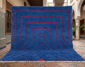 May include: A large, rectangular blue rug with a geometric red design. The rug has a thick, plush texture and a fringe along the bottom edge. The red design consists of a series of connected rectangles and lines, creating a modern, eye-catching pattern.