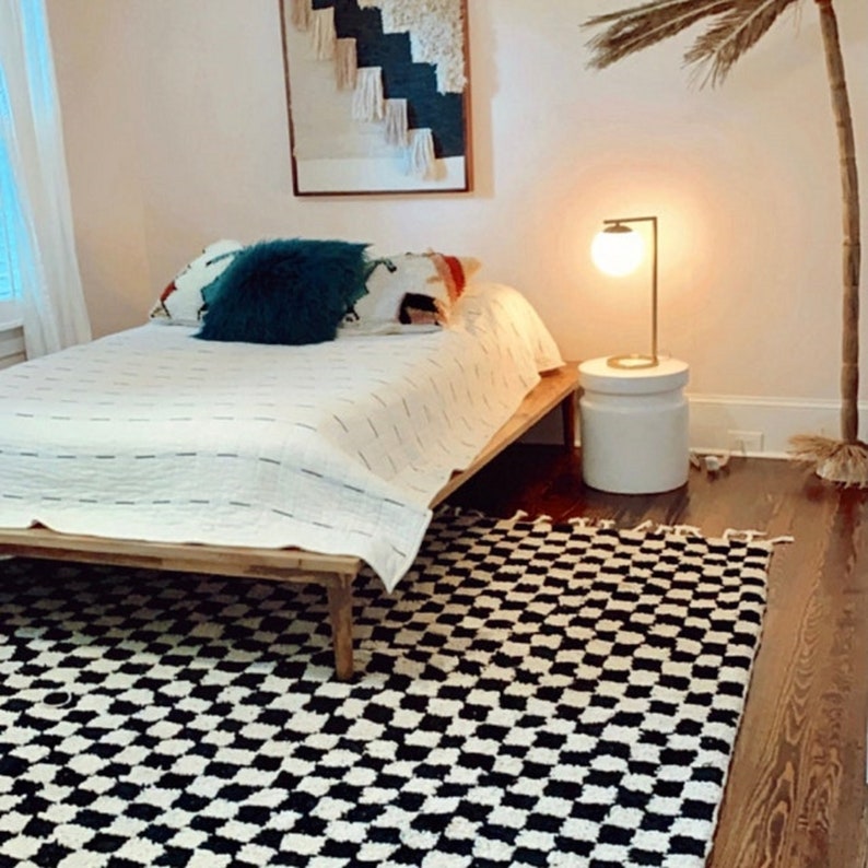 May include: A white bed with a black and white checkered rug in front of it. The bed has a wooden frame and a white duvet. There is a teal pillow on the bed and a lamp on a white side table.