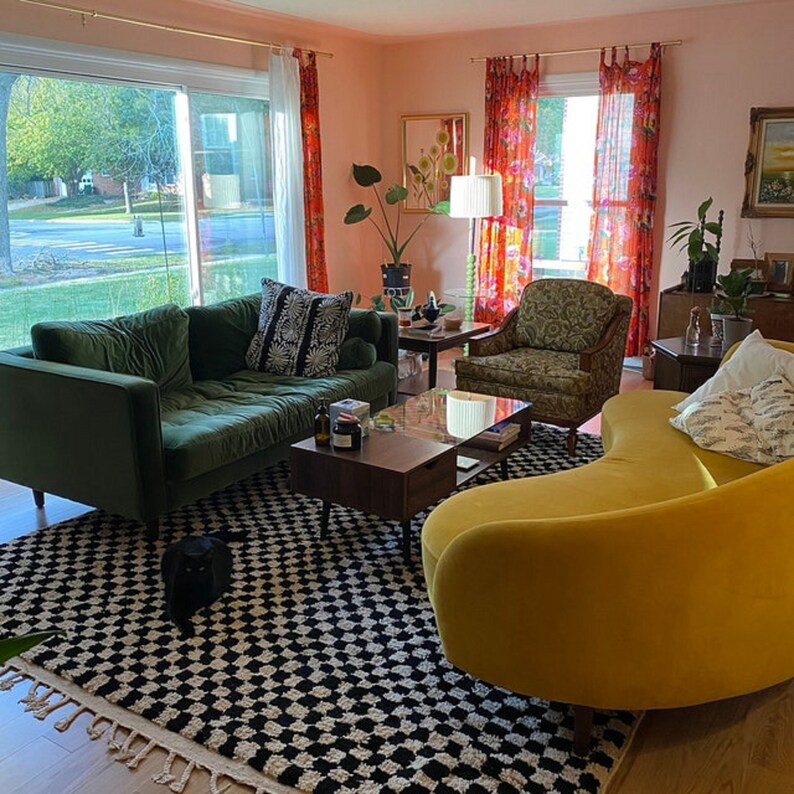 May include: A living room with a green velvet sofa, a yellow curved sofa, and a black and white checkered rug. The room has pink walls, red curtains, and a coffee table with a glass top.