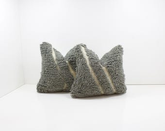 May include: Two gray, textured throw pillows with a cream-colored stripe down the center. The pillows are triangular in shape and sit on a white surface. The pillows have a soft, shaggy texture.