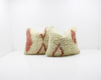 May include: Two cream-colored throw pillows with a textured, shaggy appearance. Each pillow features a geometric design in a reddish-pink hue. The pillows are placed against a white background.