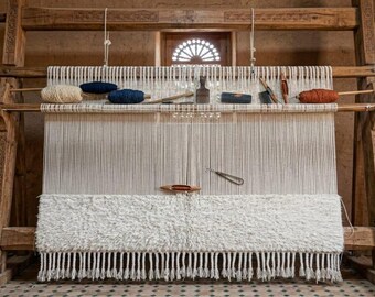May include: A wooden weaving loom with a partially completed white rug. The loom holds spools of yarn in cream, navy, and rust colors. Tools are arranged above the warp threads. The rug has a textured, fringed design.