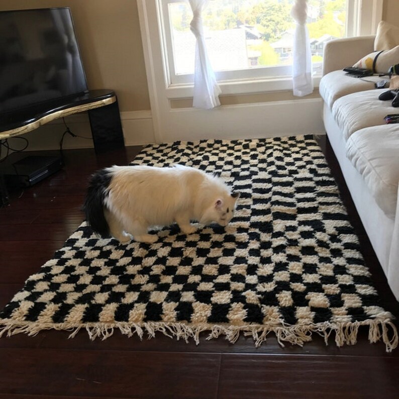 May include: A white and black checkered rug with a white cat with a black tail walking on it.