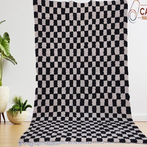 Handmade Moroccan Berber rug featuring a classic black and white checkered pattern, also known as a Checkerboard Beni Ourain rug. This plush, high-pile wool carpet is laid on a polished light oak wood floor next to a vintage wooden sideboard. The rug showcases intricate hand-knotted textures with traditional white tassels on the edges. Perfect for modern farmhouse, Scandi, or minimalist home decor, adding a cozy, bohemian touch to living rooms, bedrooms, or trendy nursery spaces.
