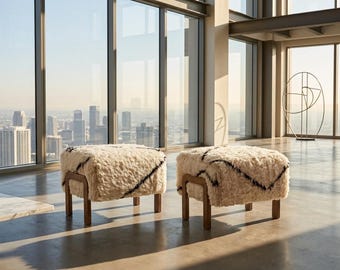 May include: Two square ottomans with cream-colored, shaggy upholstery and black zigzag patterns. The ottomans have wooden legs and are placed in a modern room with large windows overlooking a cityscape. The room has a minimalist design.