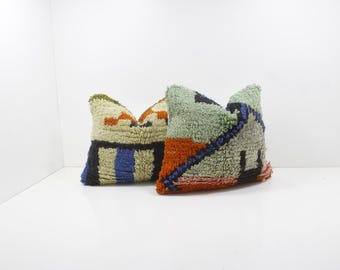 May include: Two decorative throw pillows with textured, geometric patterns. One pillow features a cream background with orange, black, and blue accents. The other has a green background with orange, black, and blue designs. Both pillows have a soft, plush appearance.