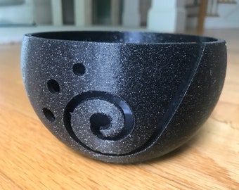 May include: A black yarn bowl with a swirl design and holes for yarn to feed through. The bowl has a speckled texture.