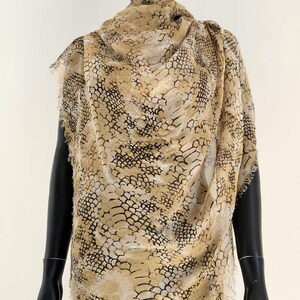 May include: A beige and black patterned scarf draped over a black mannequin. The scarf has a snakeskin-like design with frayed edges. The mannequin is set against a white background.