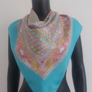 May include: A square silk scarf with a vibrant floral and paisley pattern in pink, yellow, and lavender. The scarf is displayed on a mannequin with a turquoise blue background. The scarf is a fashion accessory.