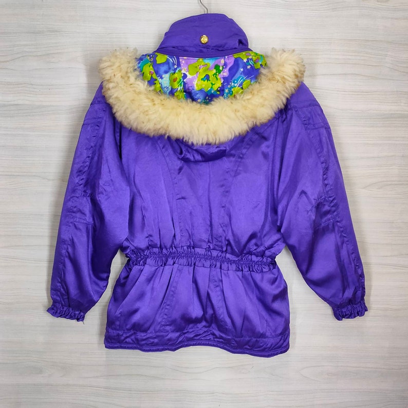 May include: A purple ski jacket with a faux fur hood lined with a floral print. The jacket has a drawstring waist and elastic cuffs.