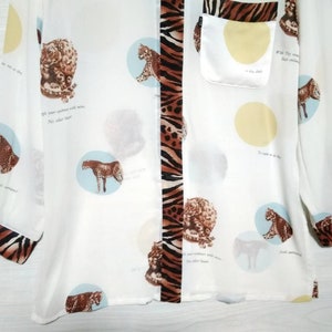 May include: White long-sleeved button-up shirt with a brown and black zebra print down the center. The shirt has a small pocket on the left side and a repeating pattern of leopards in circles throughout the fabric. The text "Wild they roam, their instincts untamed" is printed on the shirt.