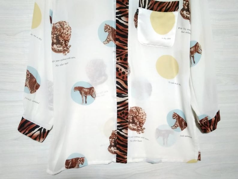 May include: White long-sleeved button-up shirt with a brown and black zebra print down the center. The shirt has a small pocket on the left side and a repeating pattern of leopards in circles throughout the fabric. The text "Wild they roam, their instincts untamed" is printed on the shirt.