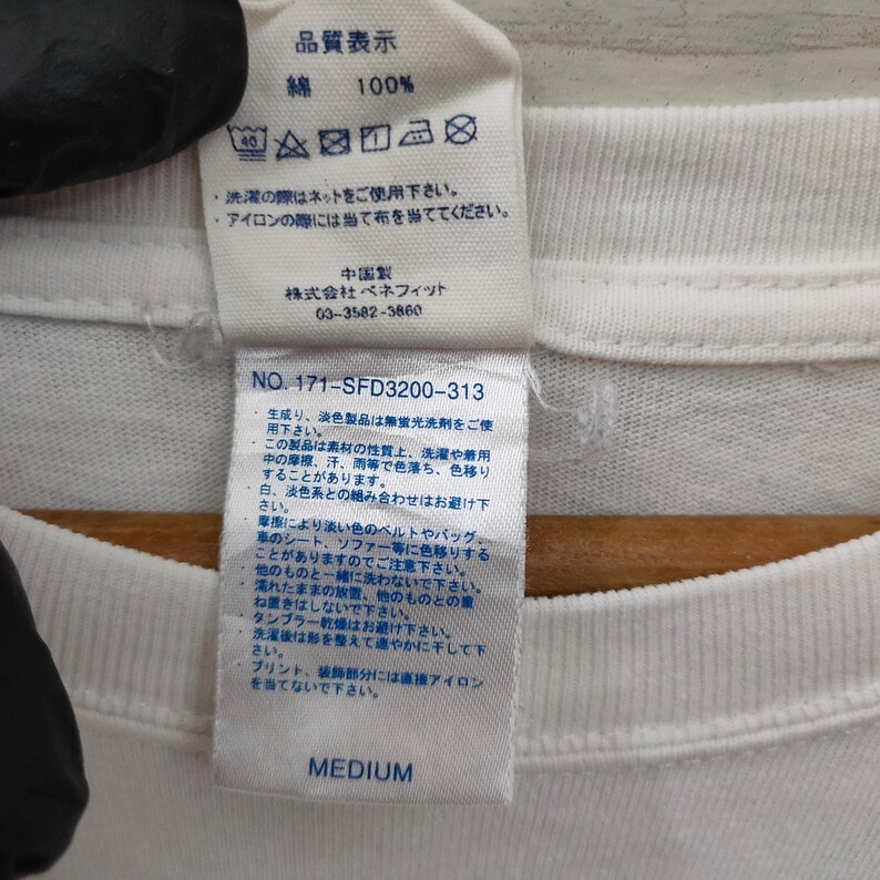 May include: Close-up of a white t-shirt tag with Japanese text and care instructions. The tag includes the text "MEDIUM" at the bottom. The tag also has washing and ironing instructions.