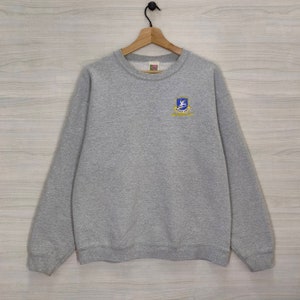 May include: A gray sweatshirt with a blue and gold embroidered patch on the left chest. The patch features a bird with wings spread and the text "SOCCER CAMP" underneath.