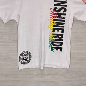 Puede incluir: Camiseta blanca con el texto "INSHINE RIDE" en negro y "Riding Wave!" en degradado arco&iacute;ris. Un gr&aacute;fico circular en la parte inferior izquierda dice "BEST SURF WAVE".
