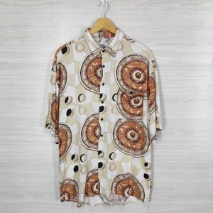 Puede incluir: Una camisa blanca de manga corta con botones y un estampado marrón y dorado de cuerpos celestes y signos del zodíaco. La camisa tiene un cuello abotonado y un bolsillo en el lado izquierdo.