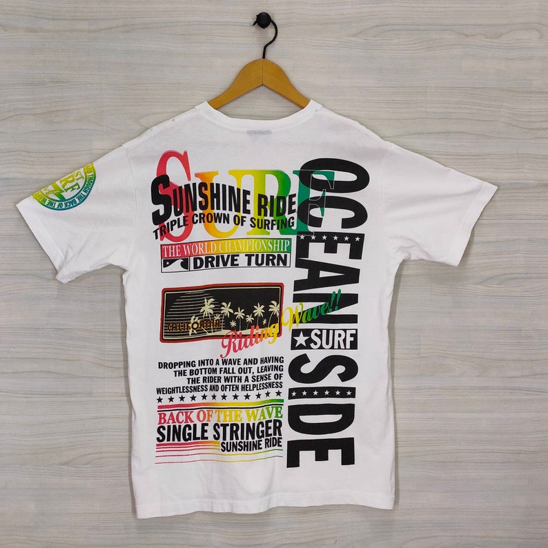Puede incluir: Camiseta blanca con un gr&aacute;fico colorido de tem&aacute;tica surf. El dise&ntilde;o incluye textos como "Sunshine Ride" y "Ocean Side". La camiseta est&aacute; colgada en una percha de madera sobre un fondo gris claro.
