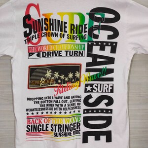 Puede incluir: Camiseta blanca con un dise&ntilde;o colorido de tem&aacute;tica surfista. El dise&ntilde;o incluye textos como "Sunshine Ride" y "Ocean Side". El gr&aacute;fico presenta palmeras y una ola, evocando un tema playero.