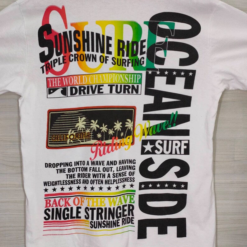 Puede incluir: Camiseta blanca con un dise&ntilde;o colorido de tem&aacute;tica surfista. El dise&ntilde;o incluye textos como "Sunshine Ride" y "Ocean Side". El gr&aacute;fico presenta palmeras y una ola, evocando un tema playero.