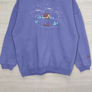 May include: A purple sweatshirt with an embroidered winter scene featuring a house, trees, and deer. The text "Cotton Club" is embroidered below the scene.