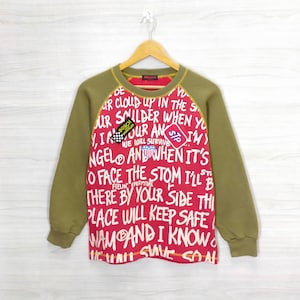 May include: A red and green long-sleeved sweatshirt with a graphic print of text and patches. The text includes "DE", "UR CLOUD UP IN THE S", "UR SMILDER WHEN YO", "VIR OUR AN SIM)", "NGELO AND WHEN IT'S", "O FACE THE STOM I'LL B", "THERE BY YOUR SIDE THI", "PLACE WILL KEEP SAFE", "WAM AND I KNOW", "MAMASIVE CON". The patches include "BOSCH SPARK PLUG" and "STP".