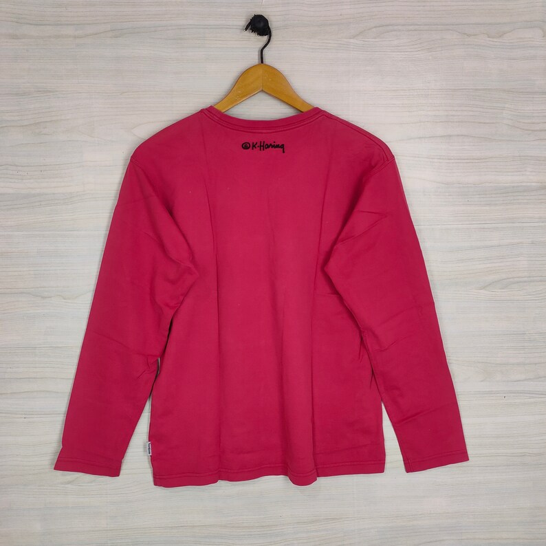 May include: A long-sleeved, red t-shirt with a black "&copy;K. Haring" logo on the back.