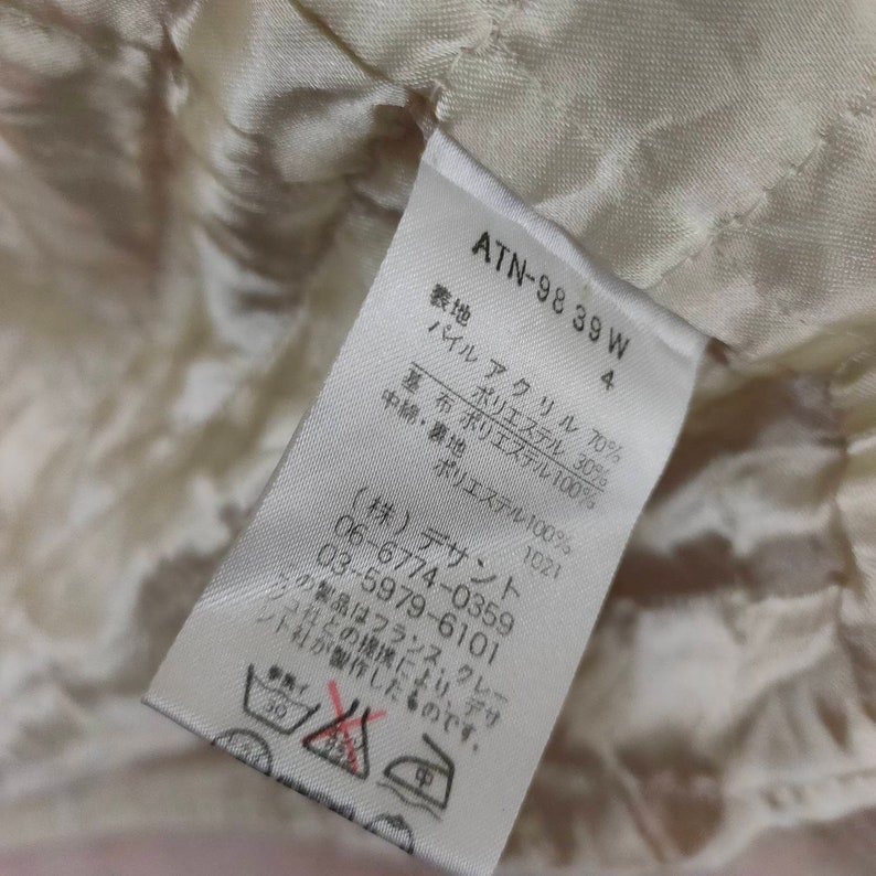 May include: White fabric label with black text detailing the composition of the garment. The label reads "ATN-98 39 W 4" and lists the fabric content as "Pile Acrylic 70%, Base Fabric Polyester 100%, Polyester 30%, Middle Layer & Lining Polyester 100%". The label also includes the company name and contact information: "(株) デサント 06-6774-0359 03-5979-6101". The label includes care instructions with symbols for washing, bleaching, drying, and ironing.