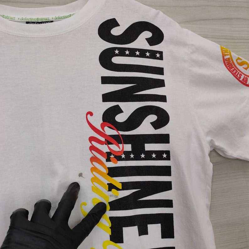 Puede incluir: Camiseta blanca con la palabra "SUNSHINE" en letras negras grandes en el lateral, con "Mirage" en un degradado de rojo, naranja y amarillo. La camiseta tiene cuello redondo y mangas cortas. El texto "defective abstract" est&aacute; impreso en el cuello.