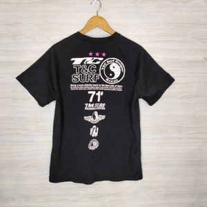 May include: Black t-shirt with a white graphic print of a yin yang symbol, text that reads "T&C Surf Designs Hawaii", and the text "Being a local at Makaha beach on the West side of Oahu". The shirt also has a white graphic print of a surfboard with wings and the text "T&C Surf Designs Hawaii" and the number "71#"