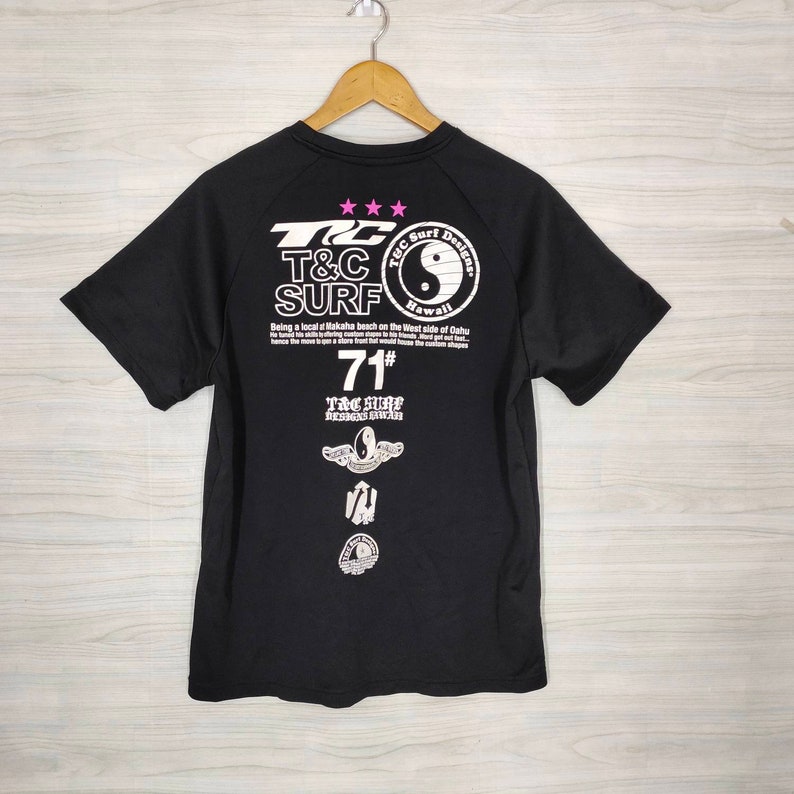 May include: Black t-shirt with a white graphic print of a yin yang symbol, text that reads "T&C Surf Designs Hawaii", and the text "Being a local at Makaha beach on the West side of Oahu". The shirt also has a white graphic print of a surfboard with wings and the text "T&C Surf Designs Hawaii" and the number "71#"