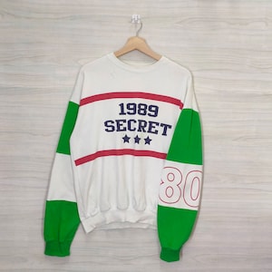 May include: White sweatshirt with green and red striped sleeves. The front of the sweatshirt has the text "1989 SECRET" in blue with three stars underneath. The right sleeve has the number "80" in white.