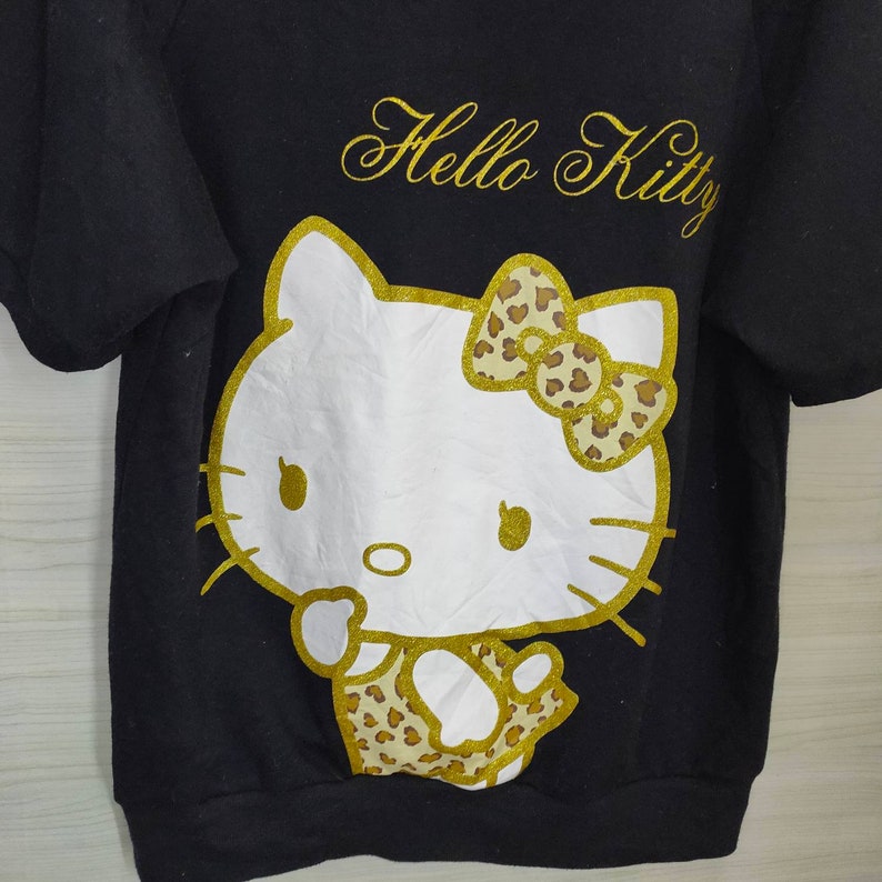May include: Black short-sleeved sweatshirt featuring a large graphic of Hello Kitty. The graphic includes a white Hello Kitty face with gold glitter outlines and a leopard print bow. The words "Hello Kitty" are written in gold script above the graphic.