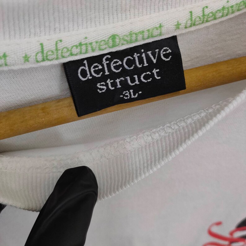 Puede incluir: Primer plano de una etiqueta de camiseta blanca con el texto "defective struct" en blanco sobre fondo negro. La etiqueta tambi&eacute;n tiene la talla "-3L". El cuello de la camisa tiene el texto "defective struct" en verde.